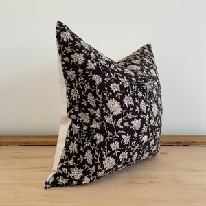 May include: Decorative pillow with a black floral pattern on a white background. The pillow features a repeating design of stylized flowers and vines. The pillow is square and appears to be made of a soft fabric.