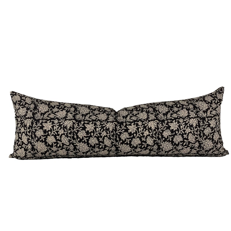 May include: A long, rectangular pillow with a black background and a cream-colored floral pattern. The pillow is designed for decorative use on a bed or sofa. The floral design is a repeating pattern of stylized flowers and leaves.