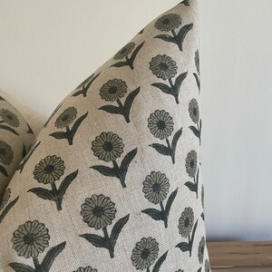May include: Close-up of a decorative pillow with a repeating floral pattern. The flowers are a muted green color with dark green stems and leaves. The background fabric is a neutral beige color.
