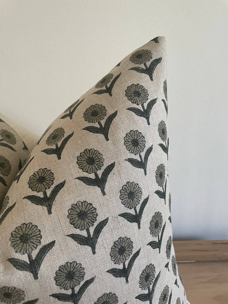 May include: Close-up of a decorative pillow with a repeating floral pattern. The flowers are a muted green color with dark green stems and leaves. The background fabric is a neutral beige color.