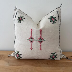 May include: Cream cushion with black edging and tassels. It features embroidered geometric patterns in pink, green, and black. The design includes a central symmetrical motif and corner accents.