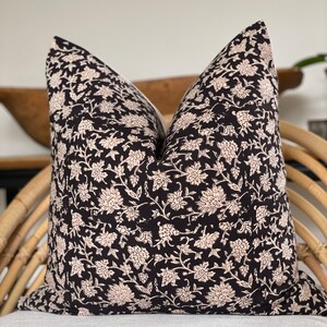 May include: A decorative pillow with a black background and a cream-colored floral pattern. The pillow is square and appears to be made of fabric. It is placed on a light-colored surface and a woven chair.