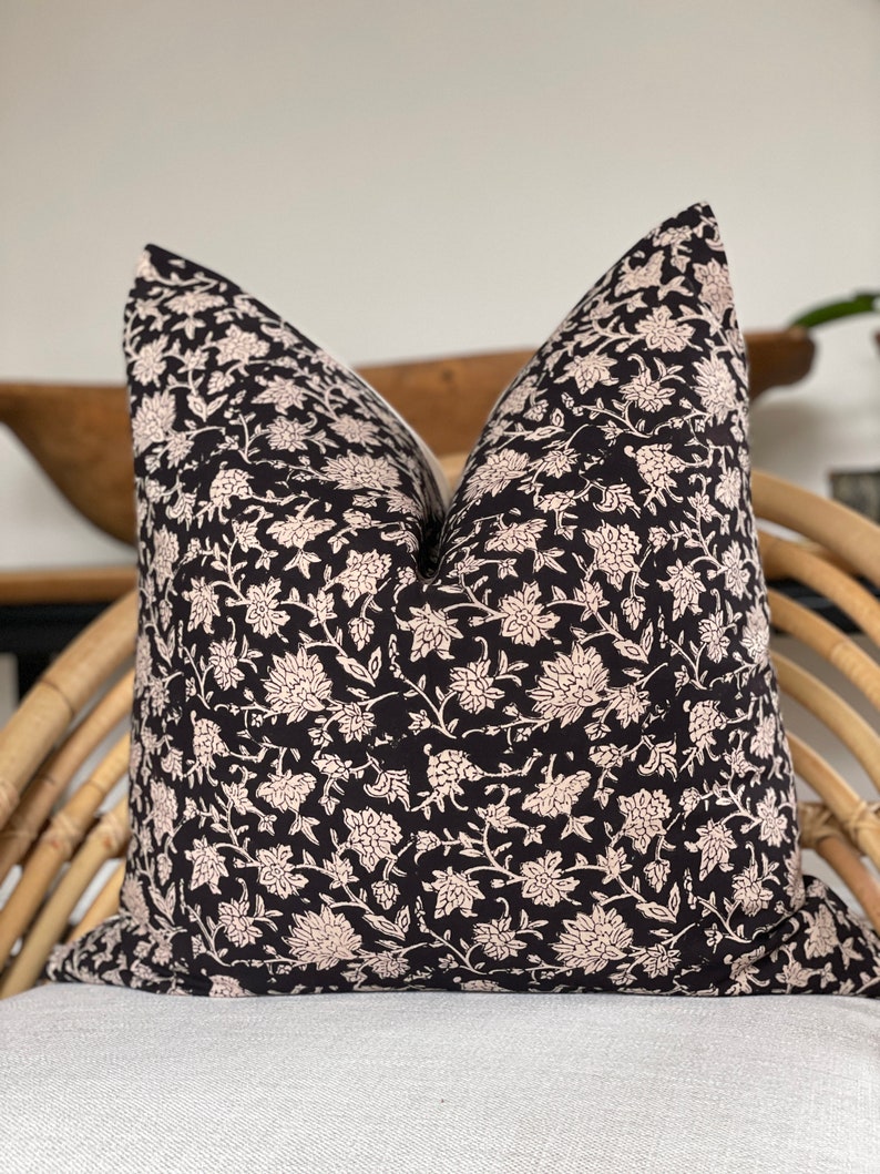 May include: A decorative pillow with a black background and a cream-colored floral pattern. The pillow is square and appears to be made of fabric. It is placed on a light-colored surface and a woven chair.