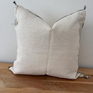 May include: Square off-white cushion with black stitching along the edges and at the corners. The cushion has a textured appearance and is on a wooden surface. Decorative accent for a sofa or bed.