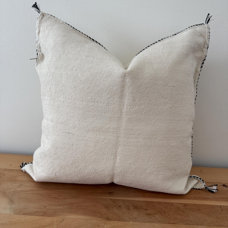 May include: Square off-white cushion with black stitching along the edges and at the corners. The cushion has a textured appearance and is on a wooden surface. Decorative accent for a sofa or bed.