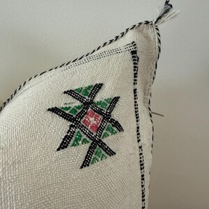 May include: Close-up of a cream-coloured decorative cushion with an embroidered geometric design. The design features black, green, and pink elements. The cushion has a black and white stitched border and a tassel.