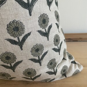 May include: A decorative pillow with a floral pattern. The pillow features a repeating design of stylized flowers in shades of gray and green on a light beige background. The pillow is made of a textured fabric.