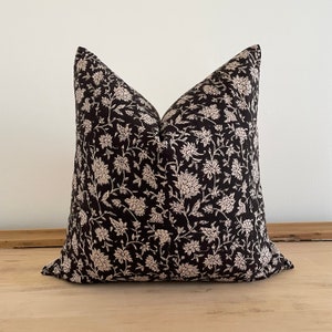 May include: A square decorative pillow with a black background and a cream-colored floral pattern. The pillow is placed on a light-colored wooden surface. The floral design features detailed flowers and leaves.