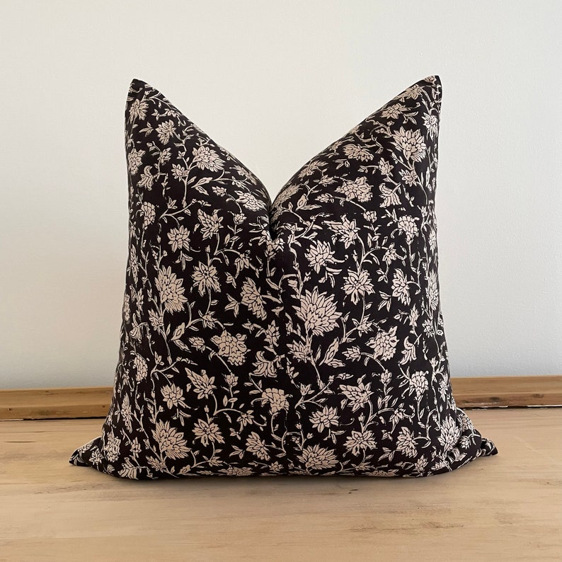 May include: A square decorative pillow with a black background and a cream-colored floral pattern. The pillow is placed on a light-colored wooden surface. The floral design features detailed flowers and leaves.