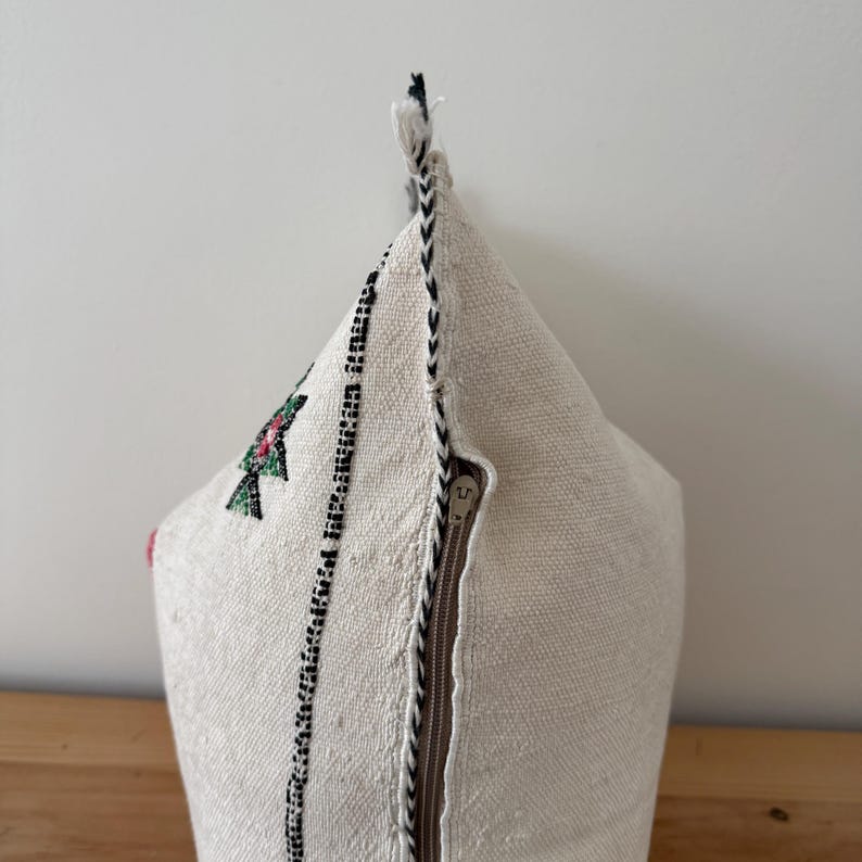 May include: A cream-coloured, triangular cushion with a decorative embroidered design. The cushion features a black and white stitched border and a brown zip. The embroidered design includes green and red floral elements.