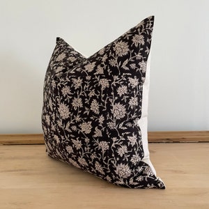 May include: A decorative throw pillow with a black floral pattern on a cream background. The pillow features a dark, intricate design of flowers and foliage, adding a touch of elegance to home decor. The pillow is square-shaped.