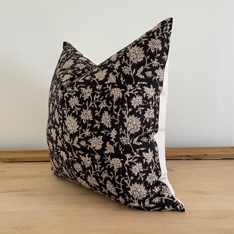 May include: A decorative throw pillow with a black floral pattern on a cream background. The pillow features a dark, intricate design of flowers and foliage, adding a touch of elegance to home decor. The pillow is square-shaped.