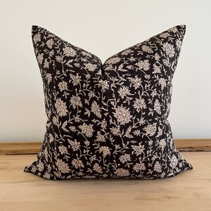 May include: A square decorative pillow with a black background and a cream-colored floral pattern. The pillow is placed on a light-colored wooden surface. The pillow's design features a repeating pattern of stylized flowers and leaves.