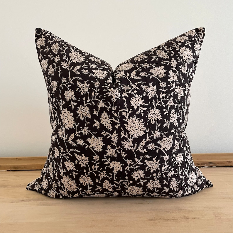May include: A square decorative pillow with a black background and a cream-colored floral pattern. The pillow is placed on a light-colored wooden surface. The pillow's design features a repeating pattern of stylized flowers and leaves.