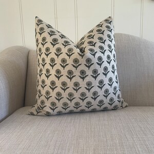 May include: A decorative throw pillow with a floral pattern. The pillow features a repeating design of dark gray flowers with stems and leaves on a light beige background. The pillow is placed on a light gray sofa.