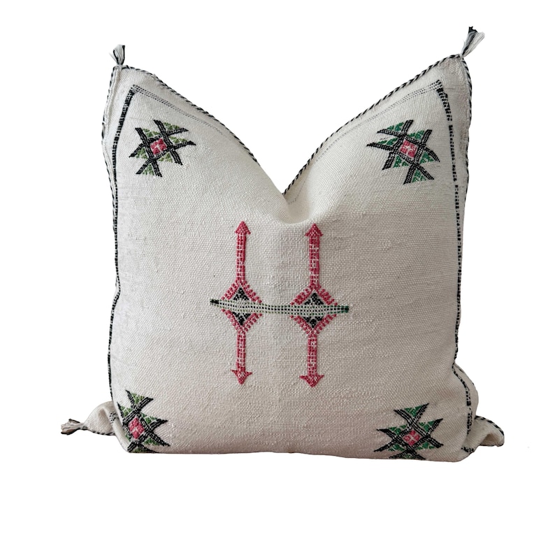 May include: Cream-coloured decorative cushion with embroidered geometric patterns in pink, green, and black. The cushion has a textured surface and a black stitched border. The design features a central vertical pattern and corner accents.