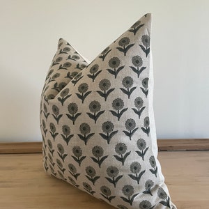 May include: A decorative throw pillow with a floral pattern. The pillow features a repeating design of dark gray flowers on a light beige background. The pillow is made of a textured fabric and is placed on a wooden surface.