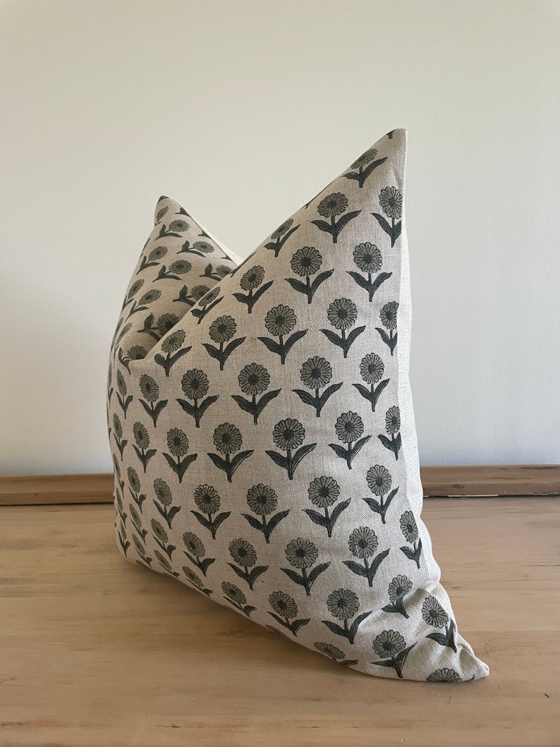 May include: A decorative throw pillow with a floral pattern. The pillow features a repeating design of dark gray flowers on a light beige background. The pillow is made of a textured fabric and is placed on a wooden surface.