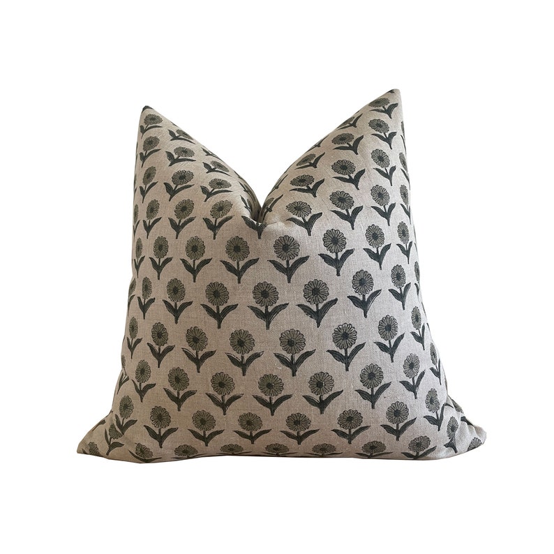 May include: A square decorative pillow with a floral pattern. The pillow features a light beige background with a repeating pattern of dark gray flowers. The flowers have a simple, stylized design, with a central circle and radiating petals. The pillow is made of a textured fabric.