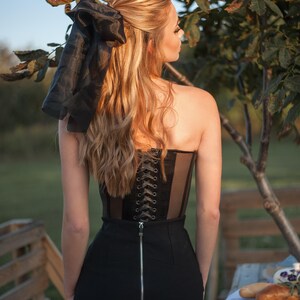 May include: A woman wearing a black corset with a lace-up back and a black skirt with a zipper. She has long blonde hair with a black ribbon tied in a bow.