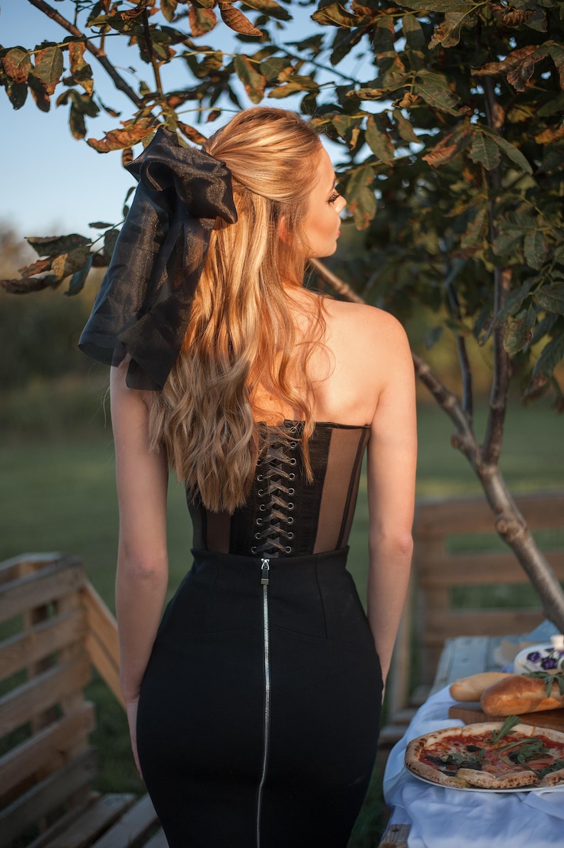 May include: A woman wearing a black corset with a lace-up back and a black skirt with a zipper. She has long blonde hair with a black ribbon tied in a bow.