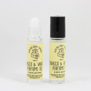 May include: Two small glass bottles of perfume oil with black roll-on tops. Both bottles have a yellow label with a cartoon bear wearing a hat and the text "Bear Hug Honey Company" and "Tobacco & Vanilla Perfume Oil". The bottles are 0.33 fl oz (9 ml).