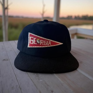 May include: A black baseball cap featuring a triangular red pennant patch on the front. The patch is embroidered with the white text GEORGIA, with a small bulldog graphic wearing a crown replacing the letter O in the center of the word.