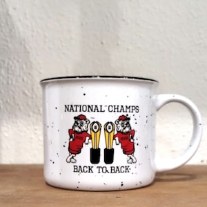 May include: White ceramic mug with black rim and speckled glaze. It features a cartoon bulldog mascot wearing a red jersey with the number 2023 on it. The bulldog is holding a gold trophy with the text "NATIONAL CHAMPS" and "BACK TO BACK" printed on the mug.