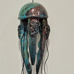 Peut inclure: Sculpture en céramique représentant une méduse, avec un dôme turquoise et bronze et des tentacules. La sculpture est suspendue par une ficelle noire, mettant en valeur une œuvre d'art unique et artisanale. La méduse a une surface texturée.