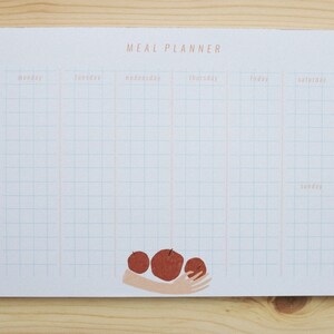 Könnte beinhalten: Ein weißer Essensplaner mit einem Rastermuster und den Wochentagen in Hellorange gedruckt. Die Worte "MEAL PLANNER" stehen oben. Eine Grafik von drei roten Äpfeln, die von einer Hand gehalten werden, befindet sich unten. Der Planer liegt auf einer Holzoberfläche.