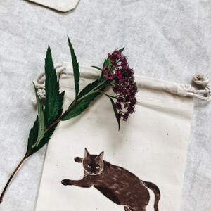 May include: A white cotton drawstring bag with a brown cat illustration and a sprig of pink flowers.