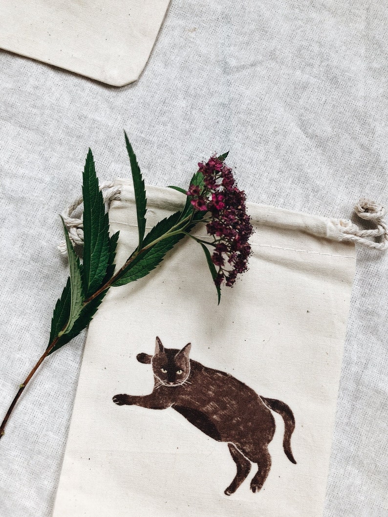 May include: A white cotton drawstring bag with a brown cat illustration and a sprig of pink flowers.