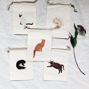 May include: Set of five small cotton drawstring bags with different cat illustrations. Each bag features a different cat breed in a simple, minimalist style. The cats are depicted in various poses, including sitting, lying down, and curled up.