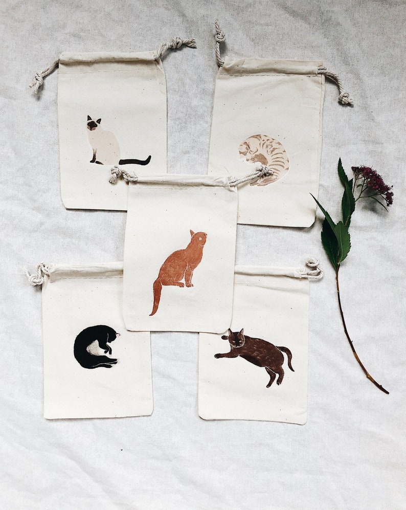 May include: Set of five small cotton drawstring bags with different cat illustrations. Each bag features a different cat breed in a simple, minimalist style. The cats are depicted in various poses, including sitting, lying down, and curled up.
