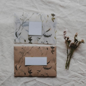 May include: Two envelopes with floral designs and blank white labels. The envelopes are on a white fabric background. A bunch of dried flowers is to the right of the envelopes.