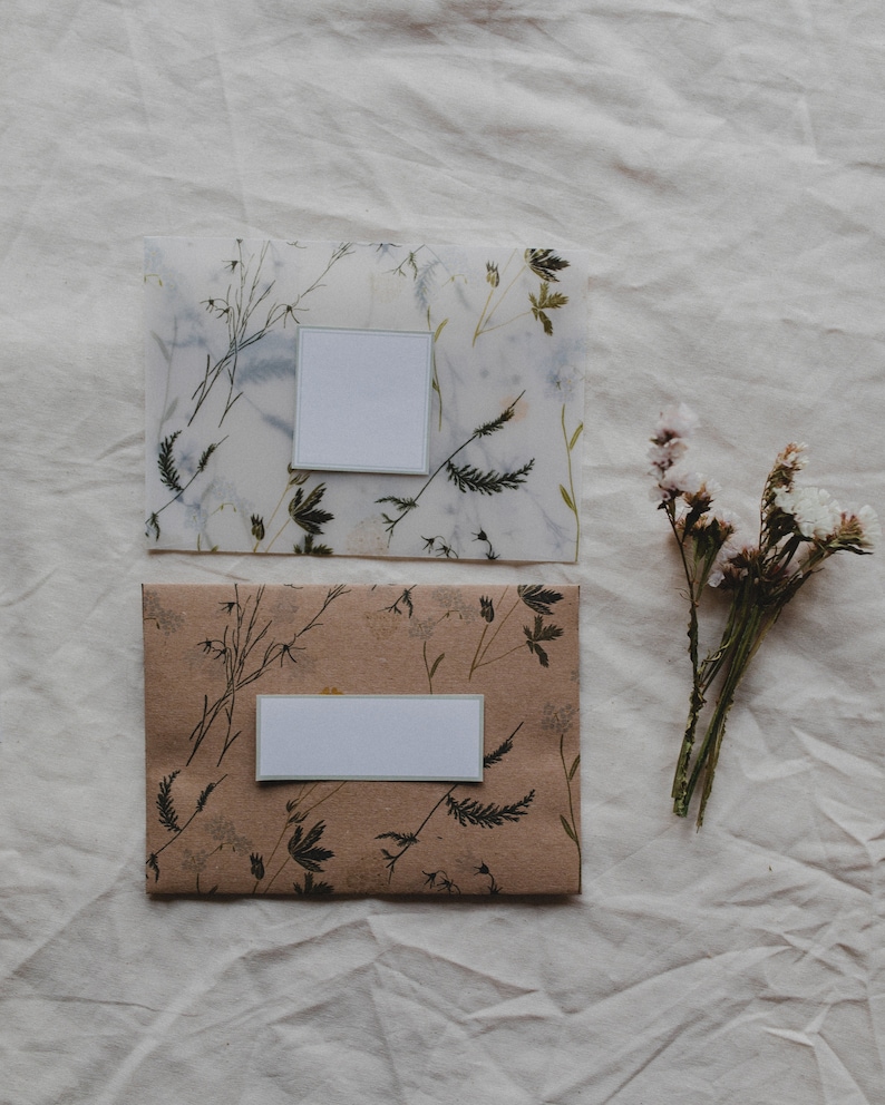 May include: Two envelopes with floral designs and blank white labels. The envelopes are on a white fabric background. A bunch of dried flowers is to the right of the envelopes.