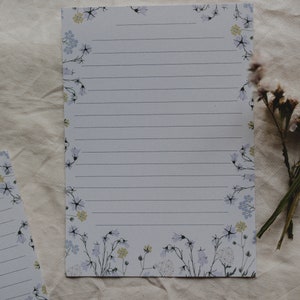 May include: A white sheet of lined paper with a floral border. The border features small blue and yellow flowers with green stems.