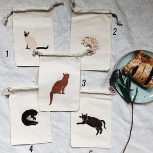 May include: Five small, white cotton drawstring bags with different cat illustrations printed on them. The bags are numbered 1 through 5.