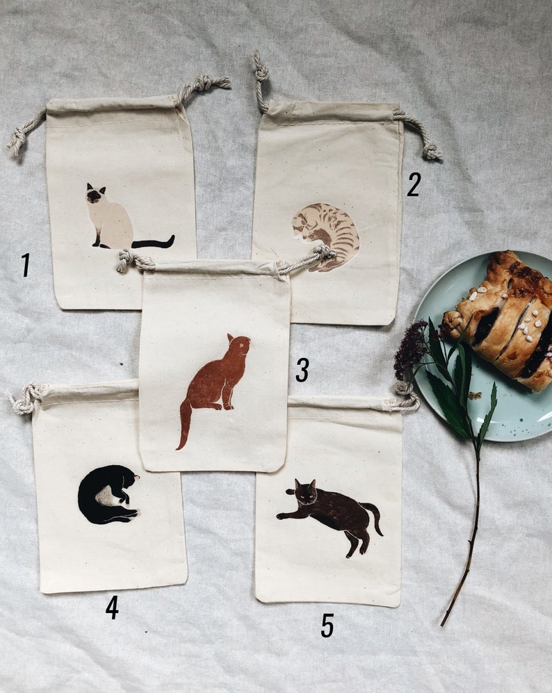 May include: Five small, white cotton drawstring bags with different cat illustrations printed on them. The bags are numbered 1 through 5.