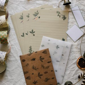Könnte beinhalten: Eine Sammlung von Schreibwaren, darunter liniertes Papier mit botanischen Illustrationen, braune und transparente Umschläge sowie dekoratives Klebeband. Eine kleine Glocke und Schnur ergänzen das Arrangement auf einem neutralen Stoff.
