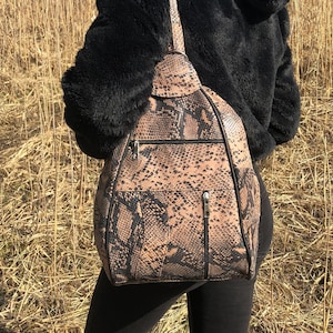 May include: A brown and black snakeskin-patterned backpack with black trim and zippers. The backpack has a single shoulder strap and a small top handle. The person in the image is wearing a black hooded jacket and black pants.