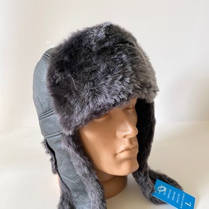 May include: A gray faux fur trapper hat with ear flaps. The hat features a faux leather exterior and a plush, furry interior and trim. A blue tag with text is attached to the hat.