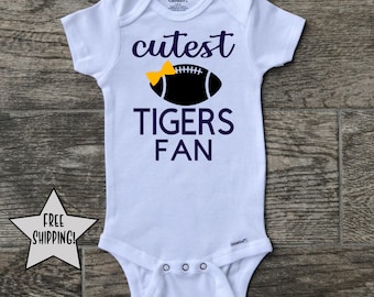 May include: White baby onesie with a black football with a yellow bow on top. The text "cutest tigers fan" is printed in purple on the onesie.
