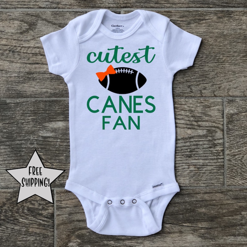 May include: White baby onesie with a green and orange design. The onesie says "cutest canes fan" with a football and a bow on top.