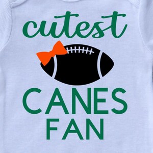 May include: White baby onesie with green text that says "cutest canes fan" and a black football with an orange bow on top.