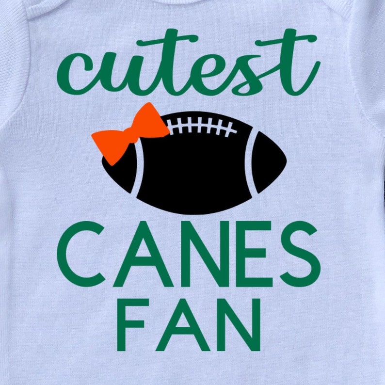 May include: White baby onesie with green text that says "cutest canes fan" and a black football with an orange bow on top.