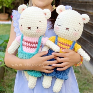 May include: Two white crocheted teddy bears wearing overalls. The bear on the left is wearing a pink and red striped shirt and teal blue overalls. The bear on the right is wearing a yellow and red striped shirt and blue overalls. www.khuccay.com