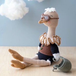 May include: A crocheted ostrich toy wearing aviator goggles and a brown and black outfit. The toy is sitting on a wooden surface with a small green crocheted hat beside it.