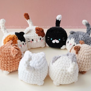 May include: A group of seven crocheted cat plushies in various colours, including white, black, brown, grey, and orange, are arranged in a circle on a white surface. The plushies have different facial expressions and are all looking in different directions.
