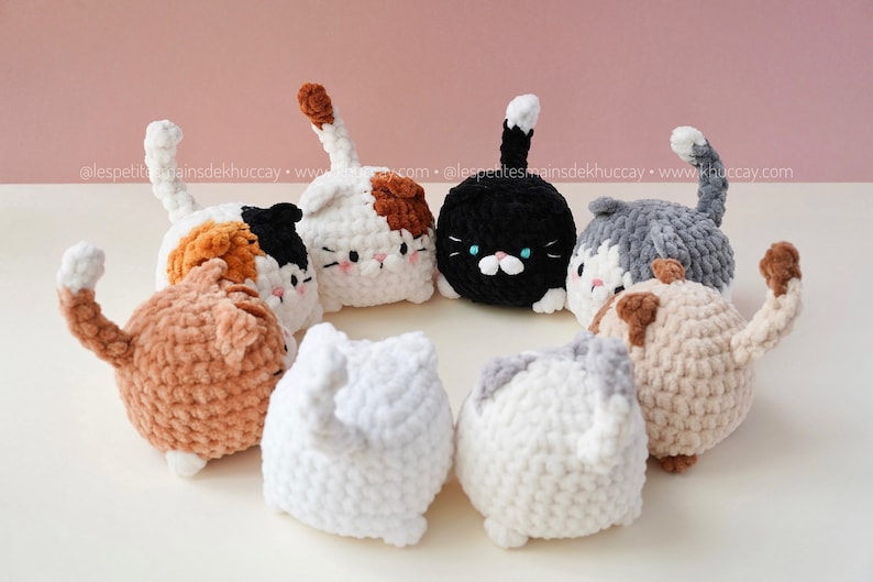 May include: A group of seven crocheted cat plushies in various colours, including white, black, brown, grey, and orange, are arranged in a circle on a white surface. The plushies have different facial expressions and are all looking in different directions.